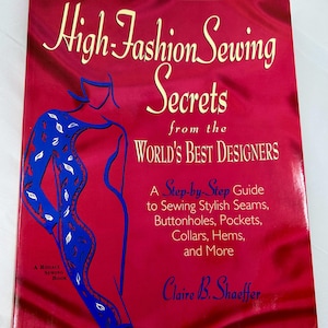 May include: A red book titled "High-Fashion Sewing Secrets from the World's Best Designers" with a blue and white illustration of a woman on the cover. The book is a step-by-step guide to sewing stylish seams, buttonholes, pockets, collars, hems, and more.