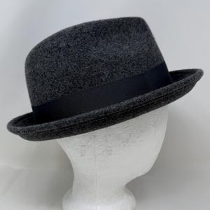 Pode incluir: Um chap&eacute;u fedora de feltro cinza-carv&atilde;o com um acabamento texturizado semelhante a l&atilde; e uma faixa de fita gorgor&atilde;o preta s&oacute;lida que circunda a copa. O chap&eacute;u possui uma aba estruturada ligeiramente levantada e uma copa vincada cl&aacute;ssica.