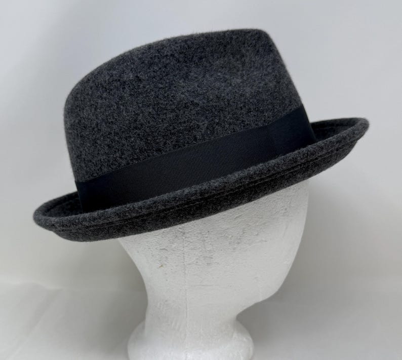 Pode incluir: Um chap&eacute;u fedora de feltro cinza-carv&atilde;o com um acabamento texturizado semelhante a l&atilde; e uma faixa de fita gorgor&atilde;o preta s&oacute;lida que circunda a copa. O chap&eacute;u possui uma aba estruturada ligeiramente levantada e uma copa vincada cl&aacute;ssica.