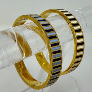 Puede incluir: Dos brazaletes dorados con un diseño a rayas. Un brazalete presenta rayas alternas de azul marino, azul claro y negro. El otro brazalete tiene rayas negras, blancas y de color beige claro. Los brazaletes se exhiben sobre un soporte transparente.