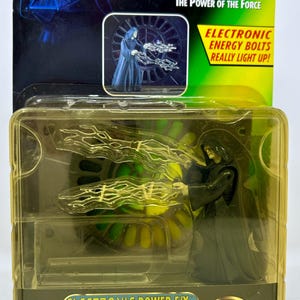May include: A Star Wars action figure of Emperor Palpatine from the Power of the Force series. The figure is dressed in black robes and has a dark side energy bolt effect. The packaging is green and yellow with the words "Electronic Power F/X" and "Galactic Empire".