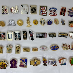 May include: A selection of Olympic pins and badges, showcasing diverse designs and logos. The pins feature gold, silver, and coloured enamel details, with text including "Atlanta 1996" and "USA". The pins are displayed on a white surface.