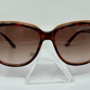 May include: Brown tortoise shell cat eye sunglasses with brown gradient lenses. The sunglasses are sitting on a clear plastic display stand.
