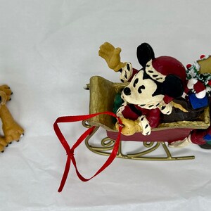 May include: A Christmas ornament featuring Mickey Mouse, Goofy, and Pluto. Mickey is wearing a red Santa hat and is in a red sleigh with a gold trim. Goofy is pulling the sleigh and Pluto is wearing reindeer antlers.