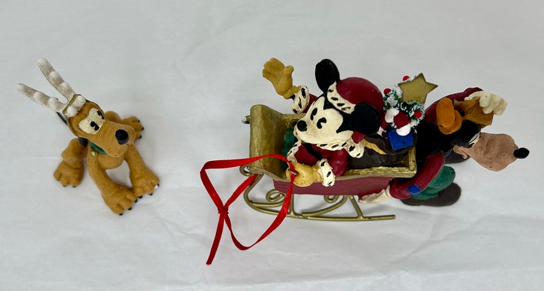 May include: A Christmas ornament featuring Mickey Mouse, Goofy, and Pluto. Mickey is wearing a red Santa hat and is in a red sleigh with a gold trim. Goofy is pulling the sleigh and Pluto is wearing reindeer antlers.