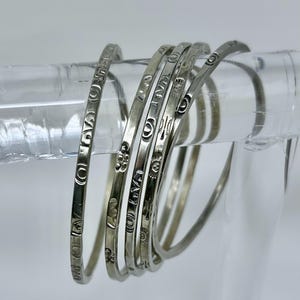 May include: Set of six silver bangles with different engraved designs. Each bangle has a unique pattern, including flowers, stars, and geometric shapes.