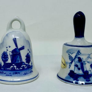 May include: Two ceramic bells with blue and white windmill designs. One bell has a loop handle, the other has a dark blue handle. Both bells feature a windmill scene with houses and trees.