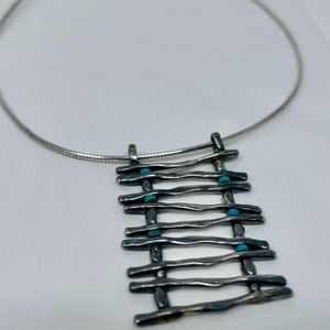 Può includere: Una collana d'argento con un ciondolo che presenta un design geometrico di barre d'argento interconnesse con pietre turchesi.