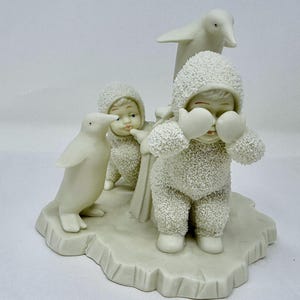 May include: A ceramic figurine depicting three children in snowsuits and two penguins on an icy base. The children are wearing mittens and hoods. The penguins are standing near the children. The figurine is painted in shades of white and off-white.