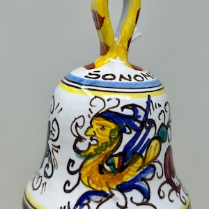May include: A white ceramic bell with a yellow, brown, and blue painted design. The bell has a handle with a yellow and brown pattern. The bell is decorated with a mythical creature and the word "SONO" in blue lettering.