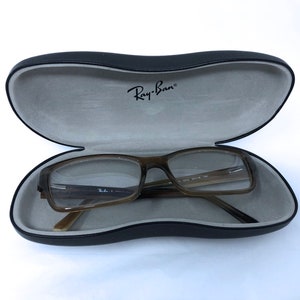 Puede incluir: Una montura de gafas rectangular marrón y negra con lentes transparentes en una funda rígida negra con interior gris. La funda tiene el texto "Ray-Ban" impreso en el interior.