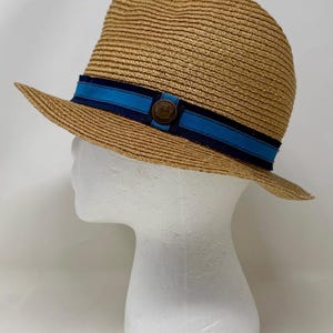 May include: A tan straw fedora hat featuring a layered hatband with a dark navy blue outer ribbon and a bright blue inner ribbon. A small, circular, dark brown metallic emblem is centered on the front of the hatband, adding a distinct detail.