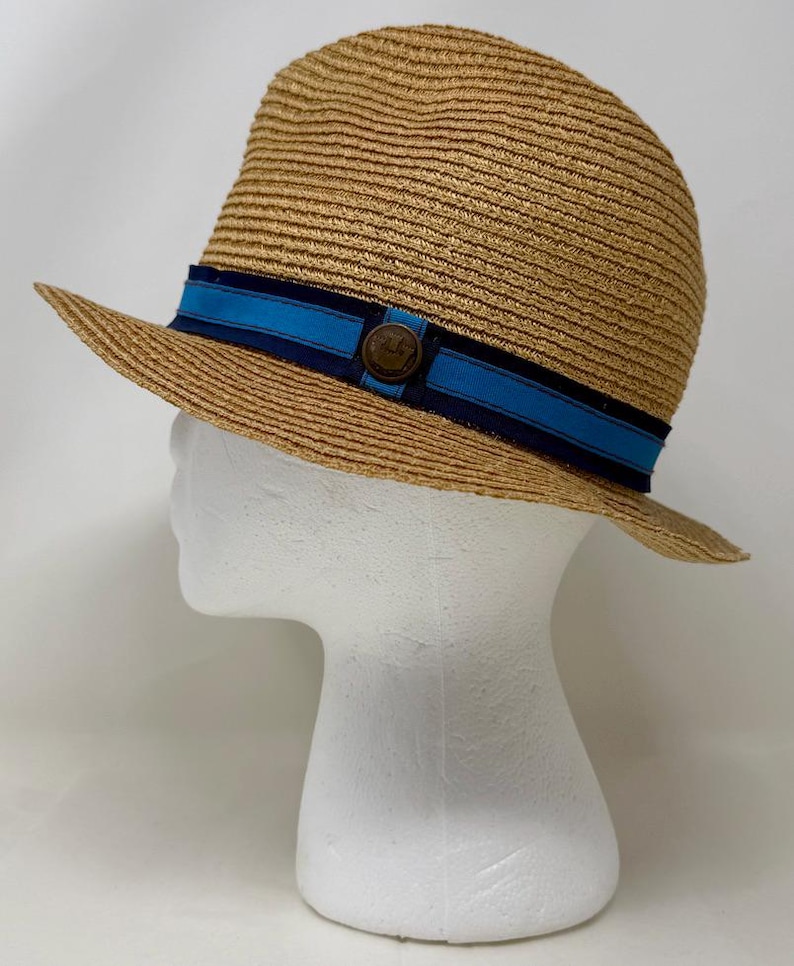 May include: A tan straw fedora hat featuring a layered hatband with a dark navy blue outer ribbon and a bright blue inner ribbon. A small, circular, dark brown metallic emblem is centered on the front of the hatband, adding a distinct detail.