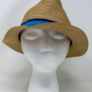 May include: A tan straw fedora hat featuring a structured crown and a wide, slightly downturned brim. The hat is accented with a horizontal band around the base of the crown, which consists of a dark navy blue stripe and a bright blue stripe.
