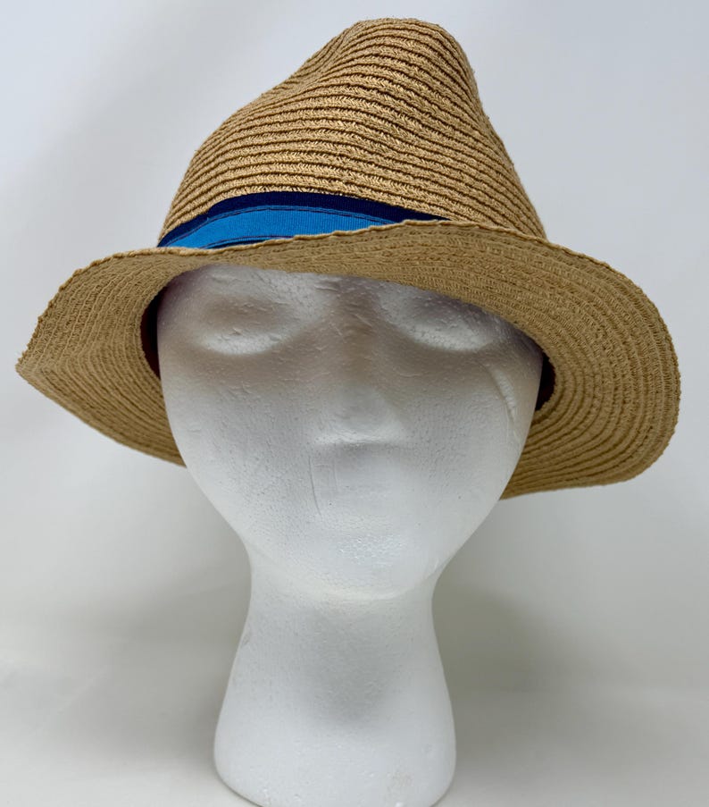 May include: A tan straw fedora hat featuring a structured crown and a wide, slightly downturned brim. The hat is accented with a horizontal band around the base of the crown, which consists of a dark navy blue stripe and a bright blue stripe.