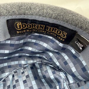 Pode incluir: O interior de um chap&eacute;u de feltro cinzento com uma etiqueta de tecido preto com bordado dourado que diz Goorin Bros Bold Hatmakers Since 1895. Uma etiqueta preta secund&aacute;ria indica o tamanho Large. O forro tem um padr&atilde;o pixelizado azul e cinzento.