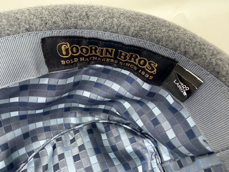 Pode incluir: O interior de um chap&eacute;u de feltro cinzento com uma etiqueta de tecido preto com bordado dourado que diz Goorin Bros Bold Hatmakers Since 1895. Uma etiqueta preta secund&aacute;ria indica o tamanho Large. O forro tem um padr&atilde;o pixelizado azul e cinzento.