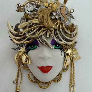 May include: A decorative face brooch with a white porcelain face, red lips, and green eyes. The brooch is adorned with gold and silver-toned metal elements, including a headpiece, star accents, and dangling chains with rhinestones. A unique piece of wearable art.