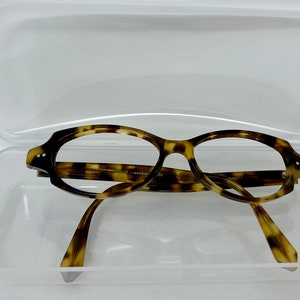 Könnte beinhalten: Schildpatt-Brille in einem transparenten Kunststoffetui. Die Brille hat eine abgerundete ovale Form und ist aus braunem und goldenem Kunststoff.