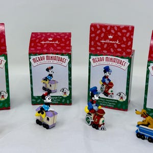 May include: Four Hallmark Merry Miniatures Christmas ornaments featuring Disney characters. Each ornament is a miniature train car with a different character, including Mickey Mouse, Goofy, and Pluto. The boxes are red with green accents.
