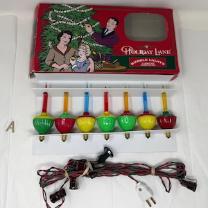 Op de afbeelding: Een set vintage Holiday Lane bubbellampjes in een rode doos met een familie-illustratie. De lampjes zijn in verschillende kleuren, waaronder groen, rood en geel. De set bevat een netsnoer en een lichtset.