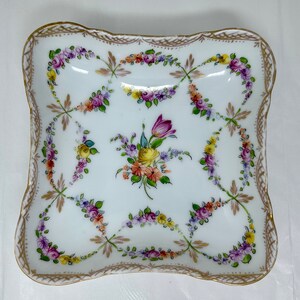 Puede incluir: Un plato cuadrado de porcelana blanca con un borde festoneado y ribete dorado. El plato está decorado con diseños florales pintados a mano en tonos de rosa, amarillo y verde.