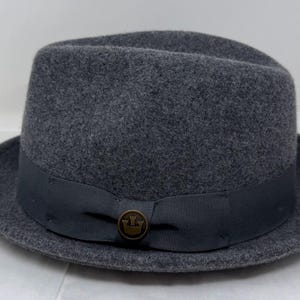 Pode incluir: Um chap&eacute;u fedora de feltro cinza-escuro com uma copa estruturada e uma aba curta ligeiramente virada para cima. Uma fita de gorgor&atilde;o cinza-escuro envolve a base da copa, acentuada com um pequeno emblema met&aacute;lico circular com um &iacute;cone de coroa.