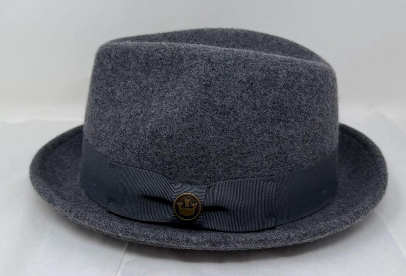 Pode incluir: Um chap&eacute;u fedora de feltro cinza-escuro com uma copa estruturada e uma aba curta ligeiramente virada para cima. Uma fita de gorgor&atilde;o cinza-escuro envolve a base da copa, acentuada com um pequeno emblema met&aacute;lico circular com um &iacute;cone de coroa.