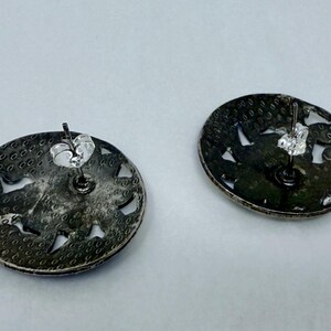 May include: Two silver-toned stud earrings. Each earring is a flat, circular disc with a textured surface and cut-out designs. The earring posts and backs are visible, indicating a secure fastening mechanism. The earrings are displayed against a light background.