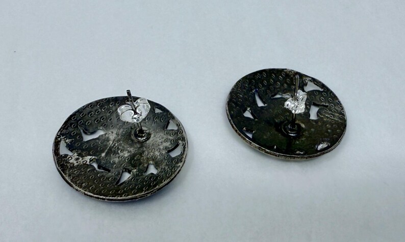 May include: Two silver-toned stud earrings. Each earring is a flat, circular disc with a textured surface and cut-out designs. The earring posts and backs are visible, indicating a secure fastening mechanism. The earrings are displayed against a light background.