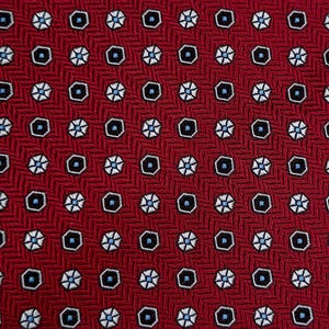 Può includere: Primo piano di un tessuto rosso con un motivo ripetuto. Il motivo presenta piccole forme di fiori bianchi a sei petali e forme geometriche nere e blu. Lo sfondo ha una sottile trama tessuta.