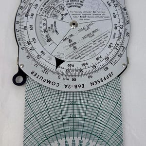 May include: A vintage Jeppesen E6B-2A flight computer with a circular slide rule and a course protractor. The device features scales for altitude, airspeed, and time, with the text "Jeppesen & Co., Denver, Colorado."
