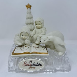 May include: A Snowbabies figurine featuring two snow-suited figures, a Christmas tree, a polar bear, and a penguin. The figures are arranged around an open book with gold accents. The base is clear and has the words "A Snowbabies Journey".