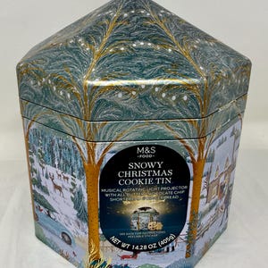 Könnte beinhalten: Sechseckige Weihnachtskeksdose mit Winterlandschaftsdesign. Die Dose zeigt eine verschneite Landschaft mit Bäumen, Hirschen und einem Haus. Das Etikett lautet "Snowy Christmas Cookie Tin" und enthält das Gewicht von 405g.