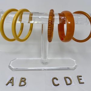 May include: A clear acrylic bracelet display stand holding five vintage bangles. The bangles are in shades of yellow and orange. Two are a solid yellow, one is carved, and two are a solid orange. The letters A, B, C, D, and E are at the base.