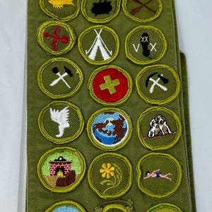 May include: A green sash with various embroidered patches. The patches feature images like a target, heart, fish, tent, and a cross. The patches are in a circular shape with a green border. The sash is made of a green fabric.