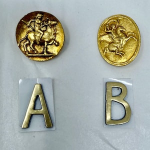 May include: Two gold-colored metal buttons with equestrian designs, one round and one oval. Below, two gold metal letters, "A" and "B", are affixed to small white rectangles. The buttons and letters appear to be vintage or antique.