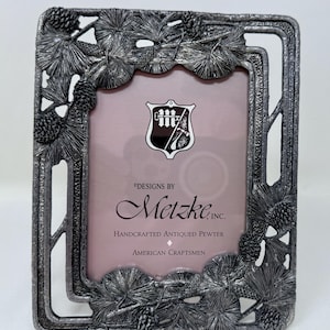 May include: Rectangular pewter picture frame with a nature-inspired design. The frame features pine needles and pine cones around the edges. The frame has the text "DESIGNS BY Metzke, INC. HANDCRAFTED ANTIQUED PEWTER AMERICAN CRAFTSMEN".