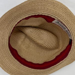 May include: A tan straw hat viewed from above, featuring a wide brim and a deep crown. The interior is lined with a dark red fabric band. A white rectangular label is stitched inside, displaying the black text Goorin Bros Bold Hatters.
