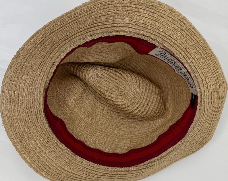 May include: A tan straw hat viewed from above, featuring a wide brim and a deep crown. The interior is lined with a dark red fabric band. A white rectangular label is stitched inside, displaying the black text Goorin Bros Bold Hatters.