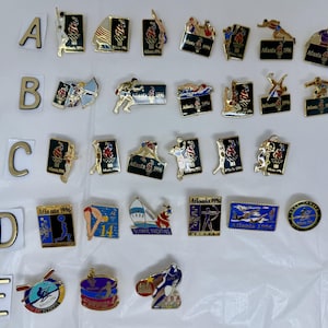 May include: A selection of vintage Atlanta 1996 Olympic pins. The pins showcase diverse sports and Olympic emblems, with a palette of gold, red, blue, and white. Several pins are inscribed with the text "Atlanta 1996".
