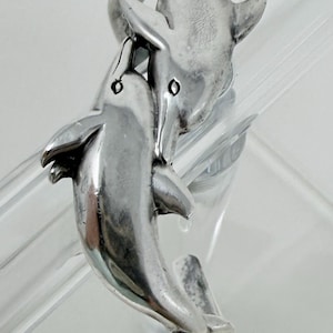 May include: A silver-toned dolphin ring featuring two dolphins in a playful embrace. The ring's design showcases the dolphins' smooth bodies and fins, with detailed eyes. The ring is a piece of jewelry, likely made of sterling silver.