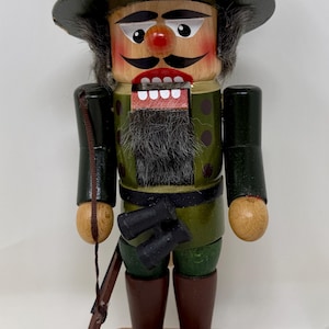 May include: Wooden nutcracker figurine of a hunter in a green outfit and hat, with a rifle and binoculars. The figurine has a red nose, black mustache, and a gray beard. The nutcracker stands on a wooden base.