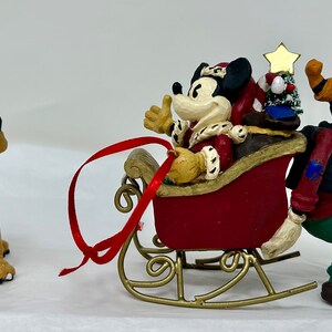 May include: A Christmas ornament featuring Mickey Mouse dressed as Santa Claus riding in a red sleigh pulled by Goofy. Pluto is wearing reindeer antlers and watching them.