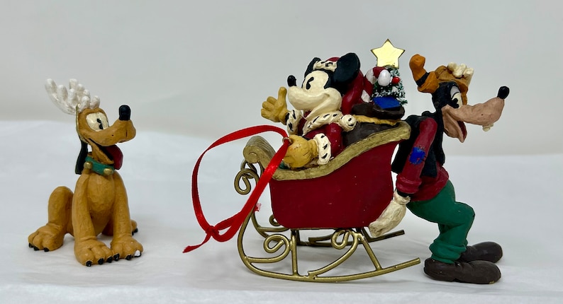 May include: A Christmas ornament featuring Mickey Mouse dressed as Santa Claus riding in a red sleigh pulled by Goofy. Pluto is wearing reindeer antlers and watching them.