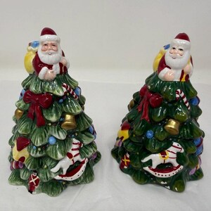 Op de afbeelding: Twee keramische kerstboombeeldjes met Santa Claus-figuren erop. De bomen zijn groen met rode strikken, gouden bellen en kleurrijke ornamenten. Elke Santa draagt een rood pak en hoed en houdt een gele zak vast.