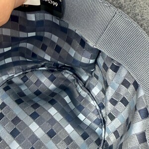 Pode incluir: O interior de um chap&eacute;u de feltro cinzento com um forro de padr&atilde;o de quadrados pixelizados em azul, cinzento e preto. Uma etiqueta de tecido preto est&aacute; presa &agrave; banda interior de gorgor&atilde;o cinzento, exibindo o texto impresso 100-5799.