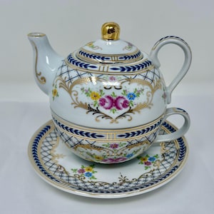 Puede incluir: Un juego de té de porcelana blanca con un diseño floral dorado y azul. La tetera tiene una tapa y un asa. La taza tiene un asa y un platillo. El juego está decorado con un patrón floral y detalles dorados.