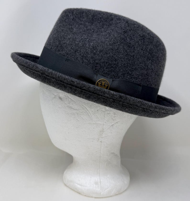 Pode incluir: Um chap&eacute;u fedora de feltro cinza-carv&atilde;o com uma faixa de fita gorgor&atilde;o preta ao redor da copa. Um pequeno emblema circular de metal em tom de bronze com um design de coroa est&aacute; fixado na lateral da faixa preta, adicionando um detalhe.