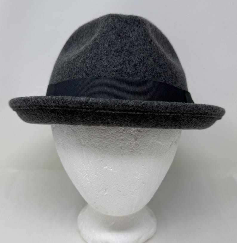 Pode incluir: Um chap&eacute;u fedora cinza-carv&atilde;o com um acabamento texturizado semelhante a feltro. O chap&eacute;u apresenta uma copa estruturada com um vinco central e uma aba curta ligeiramente levantada. Uma fita de gorgor&atilde;o preta envolve a base da copa.